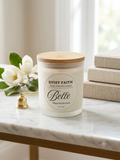A frosted glass jar candle with a light wood lid sits on a marble surface in a bright, softly lit room. The front label reads 'QUIET FAITH' and 'Belle Magnolia Blossom.