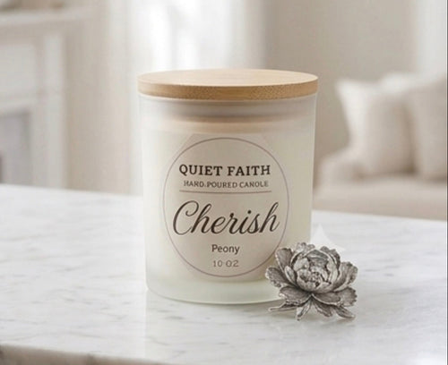 A 10 oz hard-poured candle in a frosted glass jar with a light wood lid, labeled 'Quiet Faith Cherish Peony'. It sits on a white marble surface next to a decorative gray rose in a softly blurred living room setting.