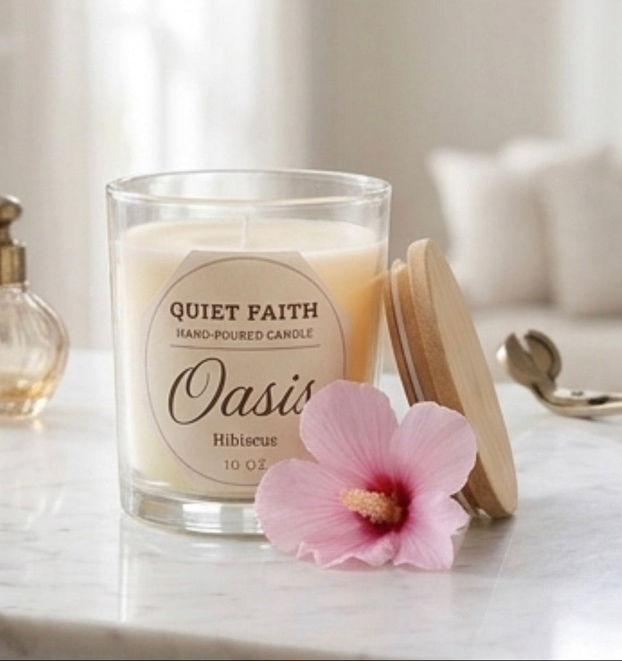 A glass jar candle labeled 'Quiet Faith' and 'Oasis' with a light wood lid, set on a marble surface with a pink hibiscus flower in front. A perfume bottle and a blurred sofa are visible in the softly lit background.