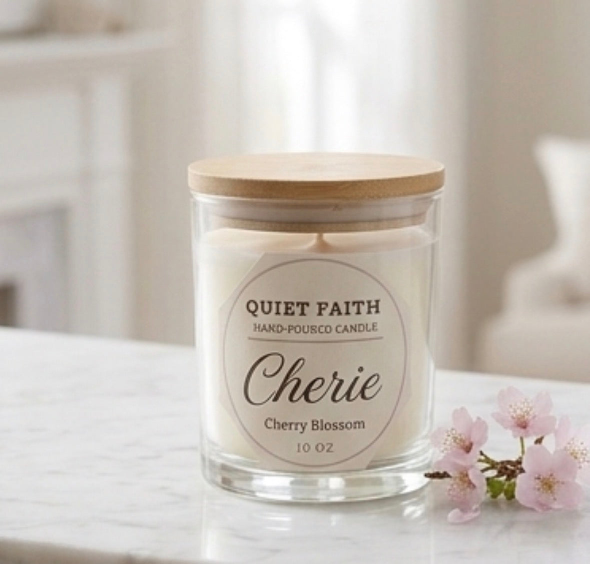 A hand-poured candle in a clear glass jar with a light wood lid, labeled 'Cherie Cherry Blossom' by Quiet Faith, sitting on a white marble surface with pink cherry blossoms nearby.